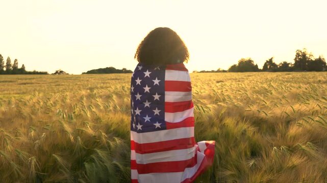 4K video clip of mixed race African American girl teenager female young woman holding and wrapped in an American US Stars and Stripes flag in a wheat field at sunset or sunrise