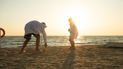 Two children play on the beach in the rays of the setting sun One child bends down to draw in the sand with a stick while the other watches. In the background is the golden ocean and the setting sun.