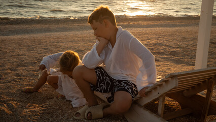 Two young children, a boy and a girl, relax on a peaceful beach, contemplating the warm golden sunset light reflecting on the sea, experiencing a quiet summer evening moment.