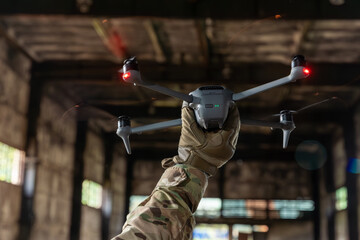 Soldier in camouflage holding a drone overhead in a dark abandoned hangar, ready for takeoff with red navigation lights on