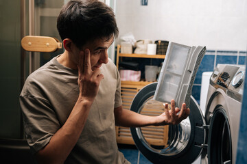 Pensive man examining dirty lint filter removed from tumble dryer, appearing concerned about large amount of lint and need for appliance cleaning and proper home maintenance.