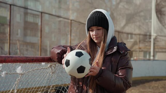 teen caucasian girl holding soccer ball near net, bundled in winter jacket and beanie, examining scuff marks while leaning on goalpost, quiet urban playground atmosphere, muted light, focused