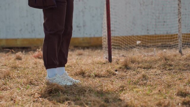 youthful player stands still on uneven field, individual in casual wear near vintage soccer goalpost outdoors, person dressed casually pauses silently beside weathered football goal in open air