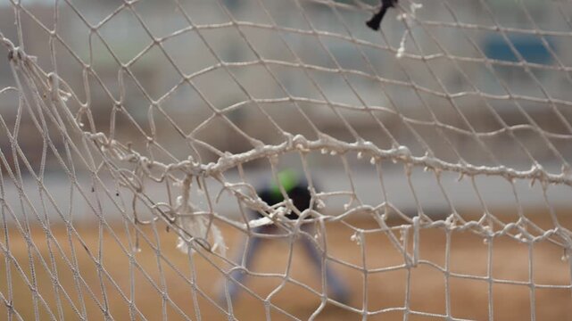 close-up view net with ball entangled and blurred goalkeeper in background, caucasian player silhouette and stadium fence visible, nylon mesh texture and tension of score moment on urban pitch