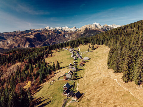 Aerial view of rustic wooden huts nestled in a valley, framed by the snow-capped Julian Alps under a clear sky, Srednja vas v Bohinju, Radovljica, Slovenia.