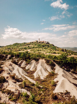 Aerial view of the ancient hilltop town with its church spire piercing the skyline, surrounded by rugged, textured terrain under a vast blue sky, Pican, Istria County, Croatia.