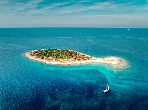 Aerial view of a small island with lush green vegetation surrounded by turquoise and deep blue waters, complemented by a lone sailboat, Fazana, Istarska zupanija, Croatia.