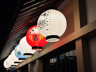A row of illuminated traditional Japanese paper lanterns (Chouchin) hanging under the eaves of a classic wooden building.