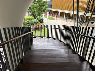 High-angle view of a modern curved outdoor wooden staircase with metal handrails, leading down to a lush green landscaped garden area on a bright sunny day.