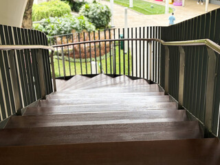 High-angle view of a modern curved outdoor wooden staircase with metal handrails, leading down to a lush green landscaped garden area on a bright sunny day.