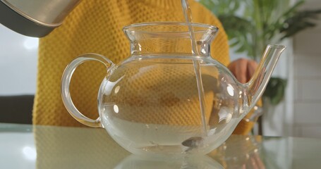 A person in a yellow sweater pouring water from a kettle into a transparent glass teapot on a table.