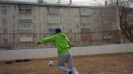 white man in bright green hoodie dribbles and volleys soccer ball across muddy urban court, beanie and grey sweatpants visible, aiming shots past small rusted goal with apartment backdrop.