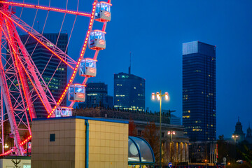 Fototapeta premium Ferris wheel at night in Warsaw with illuminated cabins, urban attraction against modern city skyline background