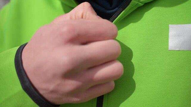 detailed shot of hand zipping jacket, practical outdoor scene with hand fastening clothing zipper detail, close view of individual fastening zipper on vibrant green outerwear in bright sunlight
