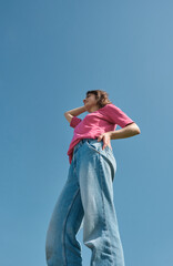 Beautiful carefree young woman with curly hair in pink t-shirt and jeans smiling and looking aside on blue sky background low angle shot.