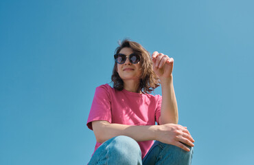Beautiful carefree young woman in sunglasses and pink t-shirt sitting and looking down at camera on blue sky background low angle shot. Copy space