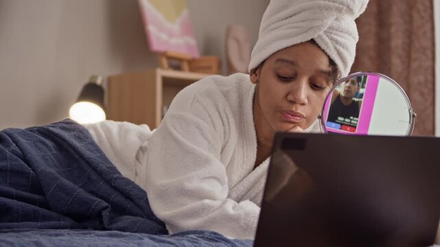 Relaxed woman with towel on head and dressed in white bathrobe lying on bed while spending time on online dating application