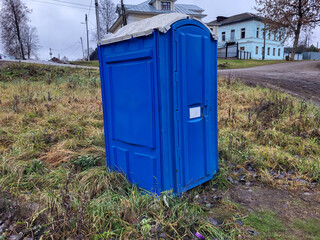 On an autumn day, a blue, single, closed plastic public toilet stall stands on the street.