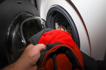 A man's hand loads a bright red fleece jacket with an open zipper into a modern white front-loading washing machine. A household process of garment care and laundry at home with an emphasis on fabric  © Олег Струс