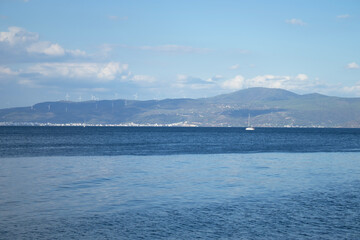 Wide view across deep blue water to hills with wind turbines and a small sailboat under soft clouds. Captured on the Marmara Sea, Turkey. Clean, commercial, sustainable-energy seascape.