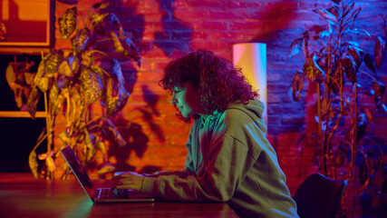Young woman typing on laptop at desk under neon light. Concept of digital work routine, remote employment, online platforms, focused execution and technology driven productivity.