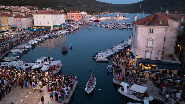 Aerial view of a vibrant harbor bustling with boats and people under the evening sky, reflecting the glow of buildings along Ulica Riva creskih kapetana, Cres, Primorsko-goranska zupanija, Croatia.