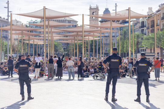 A back view of a Spanish National Police officers standing guard at a sunlit public event in Valencia. The blurred crowd and urban background emphasize a focus on public safety and security.