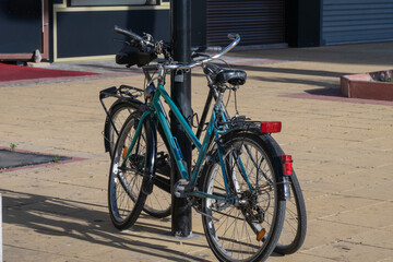 Two bicycles securely locked to a black metal pole on a sunny paved plaza, casting a sharp shadow on the yellow ground.