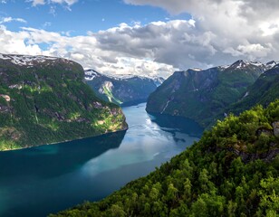 Obraz premium A breathtaking landscape depicts a deep blue fjord flanked by towering, green mountains with patches of snow under a dramatic, cloudy sky
