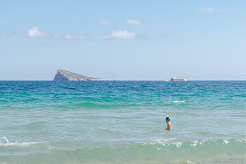Kid snorkeling with a full-face mask in turquoise waves near a sandy shore with a visible boat and an island afar in the background on a clear, bright, sunny day.