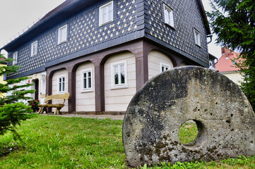 rokliwy, tradycyjny domek z ciemnym gontem na piętrze, drewnianą ławką i dużym, pamiątkowym, kamiennym kołem młyńskim. Historyczny górski kurort Kurort Jonsdorf. © Pol Badger