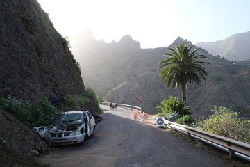 Hermigua, La Gomera, Spain : A car destroyed by a falling stones stands on mountain asphalt road. Silhouettes of hiking people on road. Canary Islands © Iwona