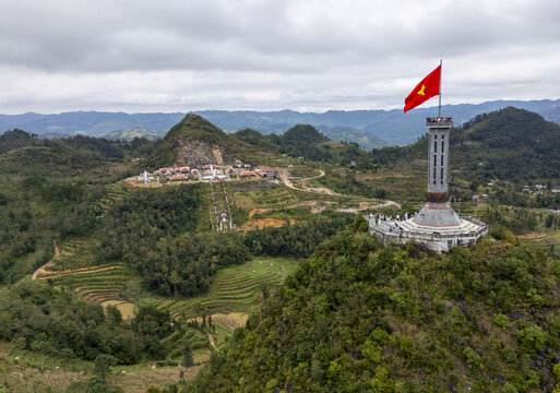 Aerial view of the vibrant Vietnamese flag atop the Lung Cu Flag Tower amid the undulating green hills, a beacon against the clouded sky, Lung Cu, Tuyen Quang, Vietnam.