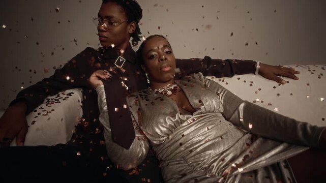 Tilt up shot of nonchalant Black woman wearing evening metallic gown reclining on sofa beside Black man while posing under falling confetti in fashion photoshoot