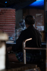Obraz premium An elderly man sitting inside a local tea shop, seen from behind, smoking and resting during the day. A candid moment reflecting everyday life and rural culture in Kerala, India.
