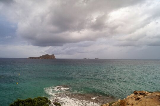Paesaggio marino visto dall'isola di ibiza con isolotto sullo sfondo cielo nuvoloso minaccioso acqua turchese e rocce in primo piano