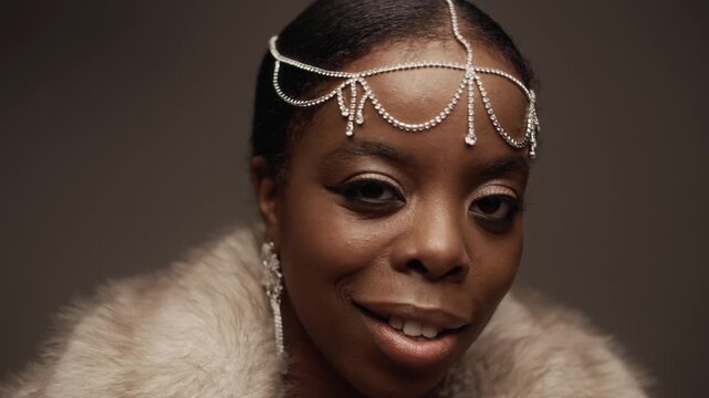 Close up shot of stylish African American woman dressed in fur coat piece and sparkling rhinestone headpiece playfully winking at camera while posing against deep brown backdrop in studio, slow motion