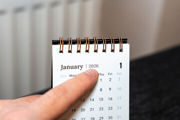 A close-up shot of a hand pointing at the January page of a 2026 calendar. The calendar highlights the first day and is set against a neutral background for a clean look.