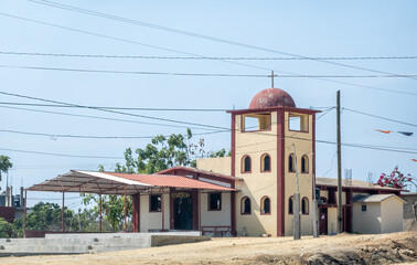 A humble Catholic chapel featuring a red dome, bell tower, and shaded outdoor area stands under power lines in a rural landscape in, Oaxaca, Mexico.