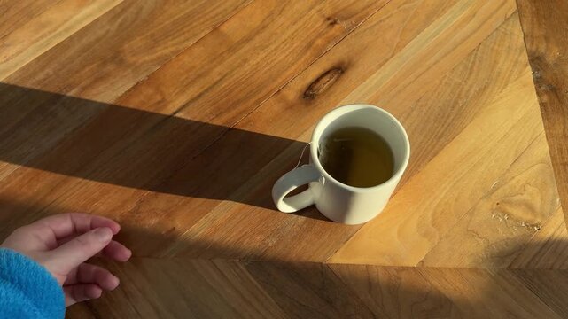 4K video of a hand dipping a tea bag into a mug of hot tea in warm natural morning light on a wooden table, cozy beverage and morning routine concept.