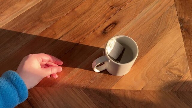 4K video of a hand placing a tea bag into a mug of hot water in warm natural morning light on a wooden table, simple tea preparation and cozy routine concept.