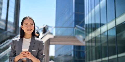 Happy young Asian business woman standing on urban street using cell phone. Smiling lady wearing...