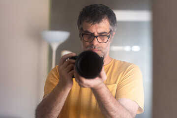 Male photographer wearing glasses and an orange t-shirt holding a professional camera taking a self-portrait in front of the mirror in his living room.