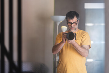 Male photographer wearing glasses and an orange t-shirt holding a professional camera taking a self-portrait in front of the mirror in his living room.