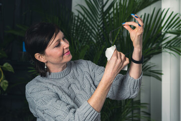 A woman is holding a pair of glasses and wiping them with a cloth. She is smiling and she is in a relaxed and comfortable mood