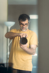 Male photographer wearing glasses and an orange t-shirt holding a professional camera taking a self-portrait in front of the mirror in his living room.