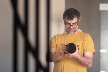 Male photographer wearing glasses and an orange t-shirt holding a professional camera taking a self-portrait in front of the mirror in his living room.
