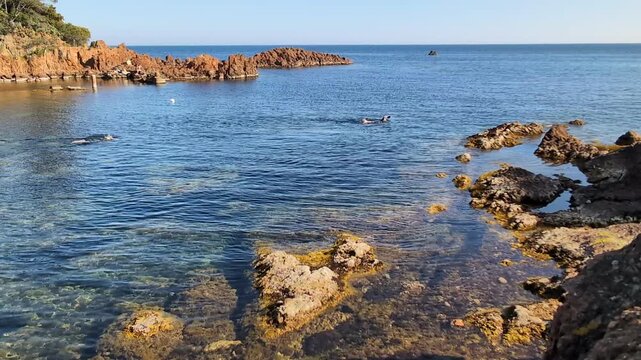 La mer est calme et tranquille, avec un fond rocailleux et des c&ocirc;tes rocheuses. On aper&ccedil;oit deux nageurs.