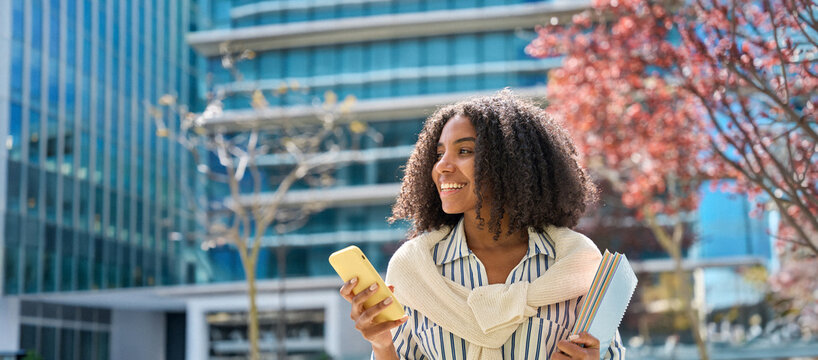 Smiling happy African teen girl student holding cellphone looking away with smartphone technology in hand walking in college park outside using apps on mobile phone, authentic shot. Banner for ads