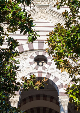 Striped marble arch and circular window of Famedio chapel at Monumental Cemetery in Milan, Italy, framed by lush green tree branches in sunlight.